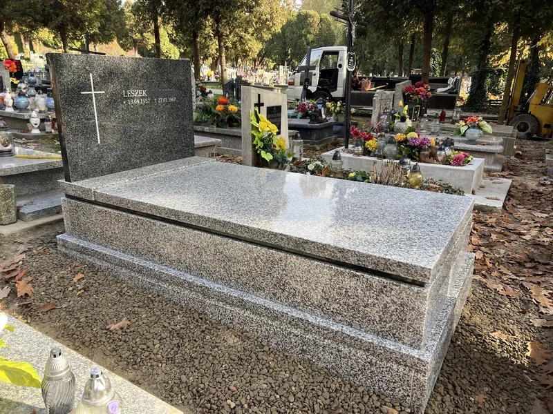 Panoramiczne ujęcie grobowca Tarn z czarną stelą Impala z jesiennymi liśćmi na płycie i ruchem cmentarnym w tle, Kraków, Kamileo.