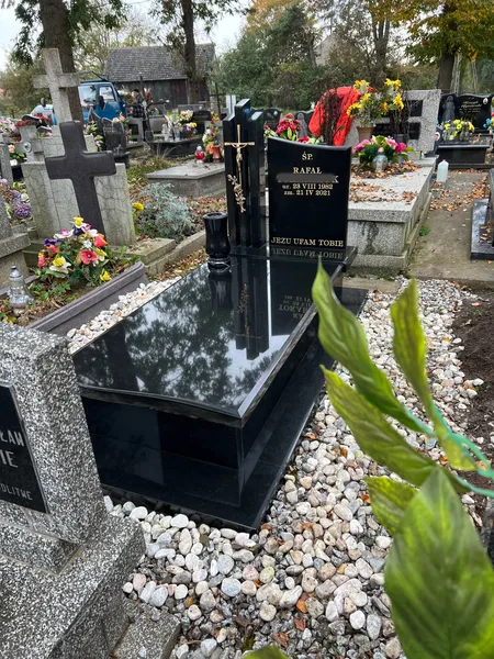Czarny nagrobek z granitu Monumental z tablicą inskrypcyjną i białym otoczakiem, ujęcie skośne przez roślinność, cmentarz Kraków, Kamileo.