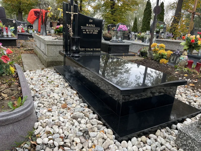 Czarny nagrobek z granitu Monumental z pozłacanym krzyżem i tablicą inskrypcyjną, obramowanie z białego otoczaka, cmentarz Kraków, Kamileo.